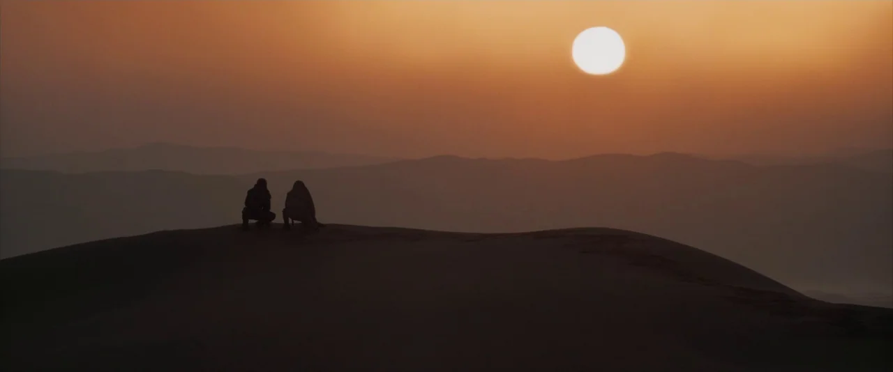 Dune: Part 2 (2024) movie still: wide — Two figures are positioned on the peak of a massive, hazy desert sand dune. They sit side-by-side as they…