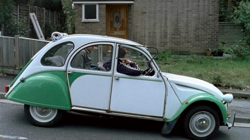 Dead Man’s Shoes (2004) movie still: wide — A green and white vintage compact car drives past a suburban brick house. Two men sit inside the vehicle…