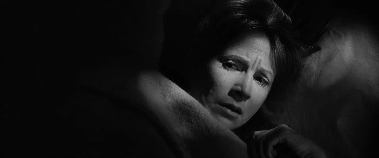 The Haunting (1963) movie still: closeup — A dark, dimly lit bedroom provides the setting for a person resting in bed. A woman lies on a pillow with…