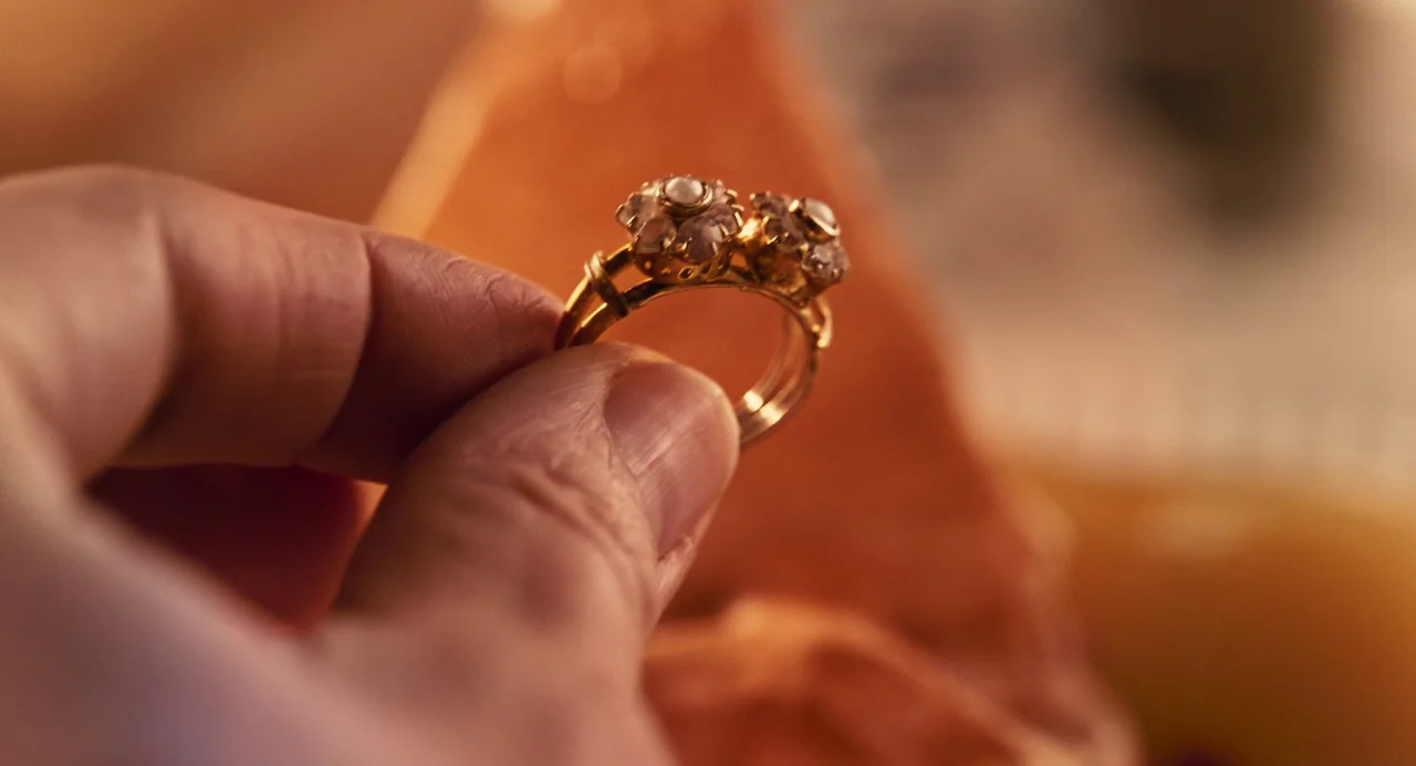 The Taste of Things (2023) movie still: closeup — An ornate gold ring with small decorative stones is held between two fingers. The background is a…