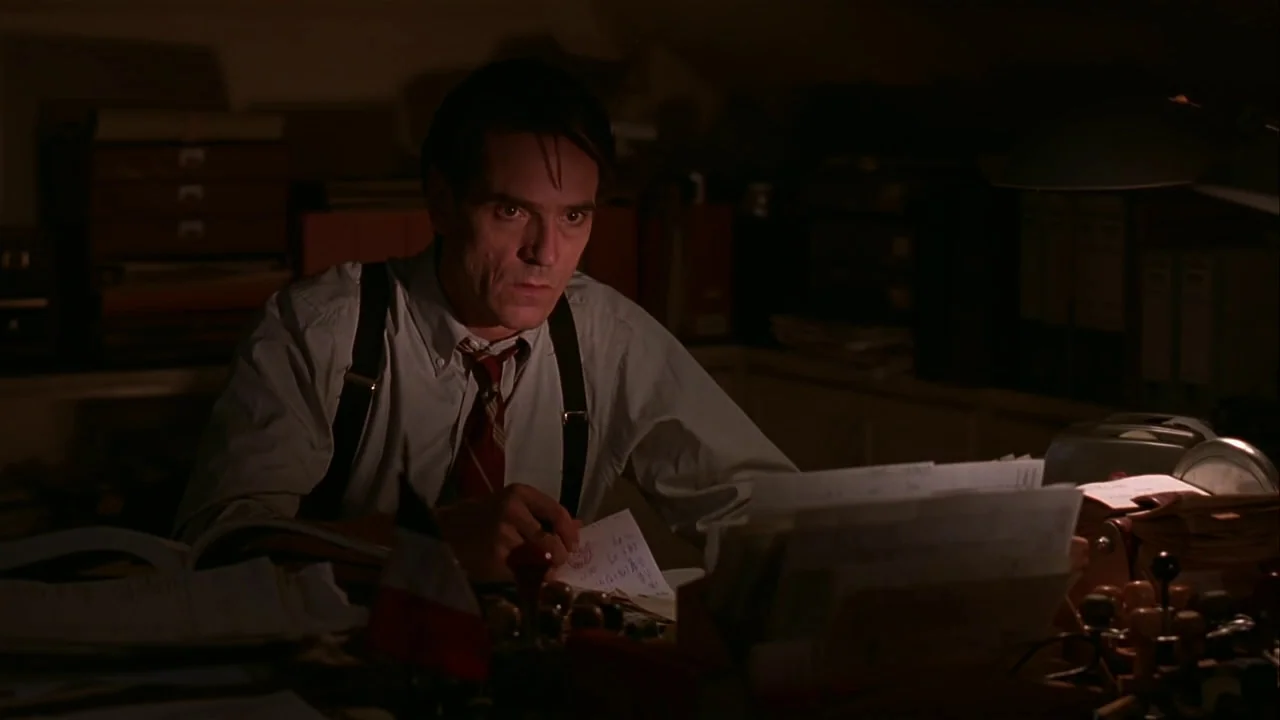 M. Butterfly (1993) movie still: medium — A dimly lit room filled with stacks of paperwork and office supplies at night. A middle-aged man works at…