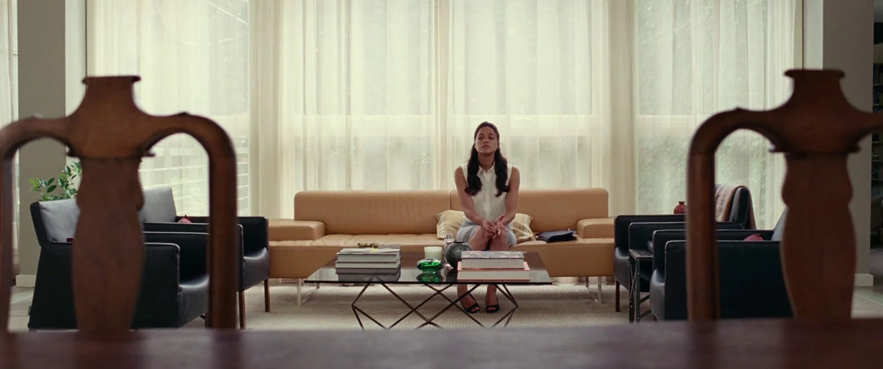 Widows (2018) movie still: wide — A modern living room features a tan leather sofa against a floor-to-ceiling curtain wall. A woman sits centered on…