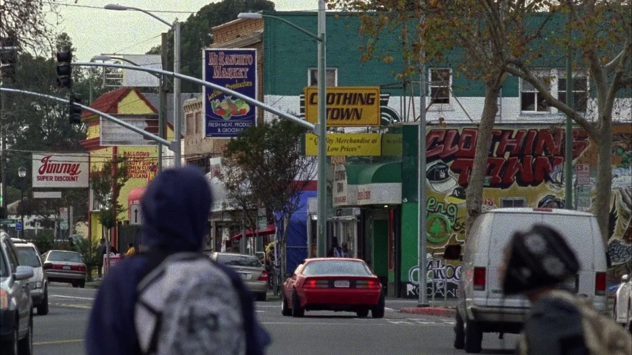 Fruitvale Station (2013) movie still: wide — A suburban street lined with colorful small businesses and commercial signs featuring painted murals.…
