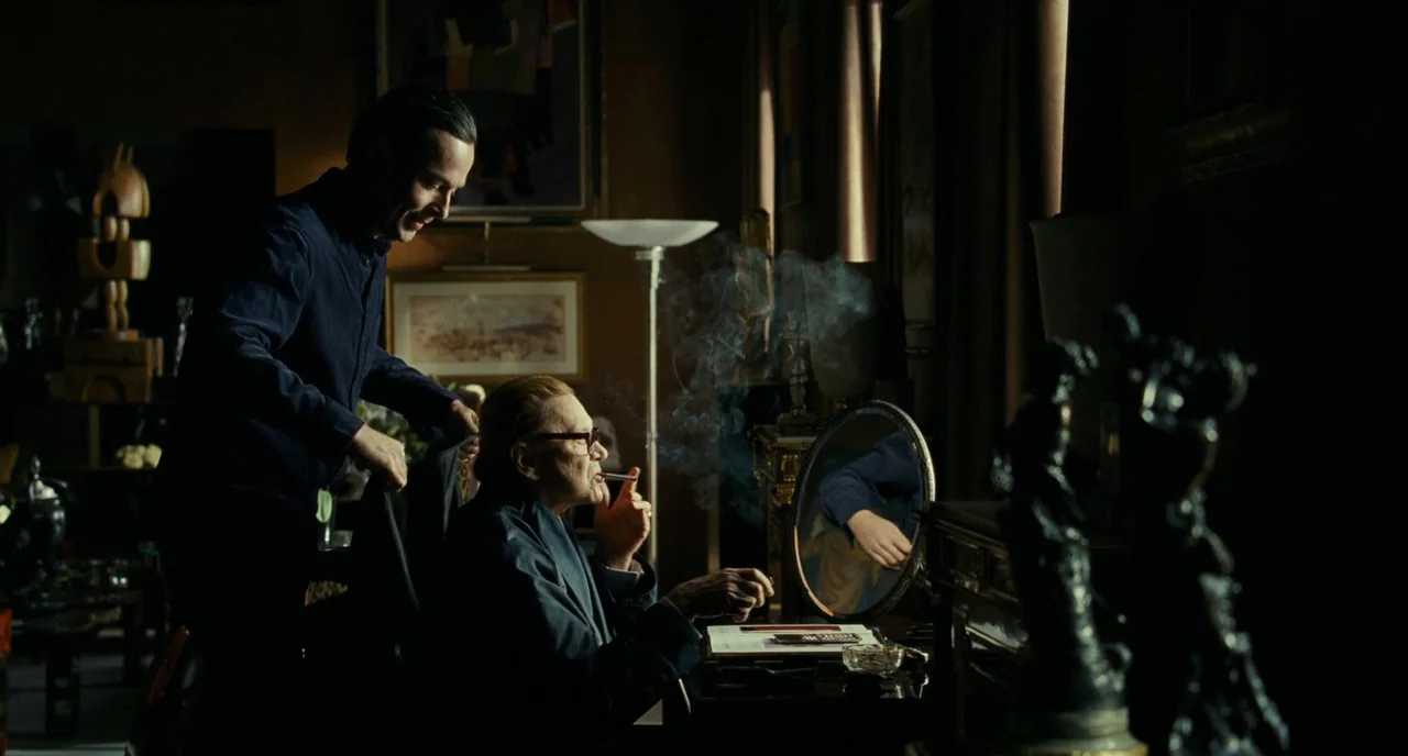Saint Laurent (2014) movie still: medium — A dimly lit room contains numerous art objects and a vanity with a mirror. A younger man smiles while…