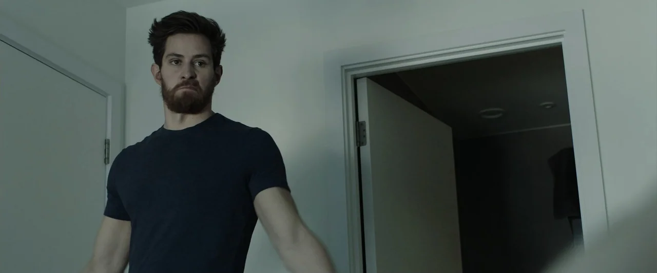 The Thorn On His Side (2014) movie still: medium — A sparse modern bedroom with white walls and an open doorway leading to darkness. A man in a blue…