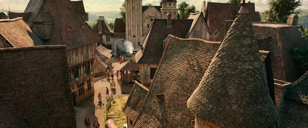 Beauty and the Beast (2017) movie still: establishing — A dense medieval village with timber-framed buildings and steep shingled roofs lines a…
