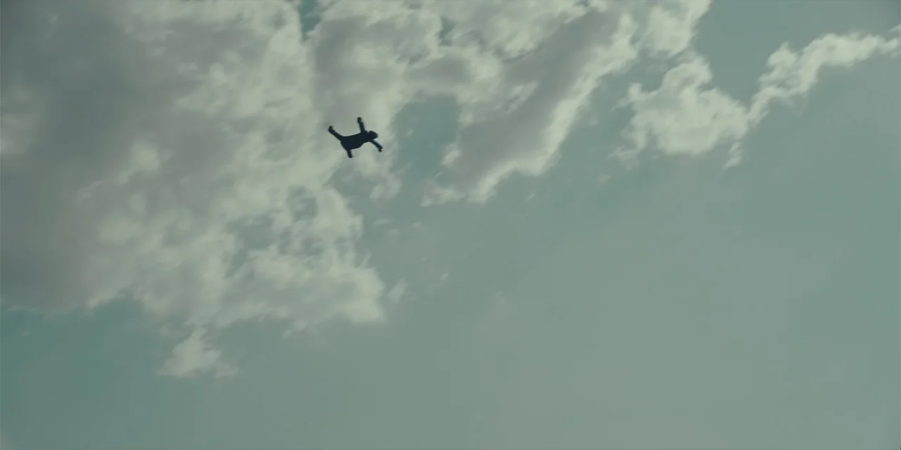 Fall (2022) movie still: establishing — A vast, open sky is filled with scattered white clouds during the daytime. A single human figure in a…