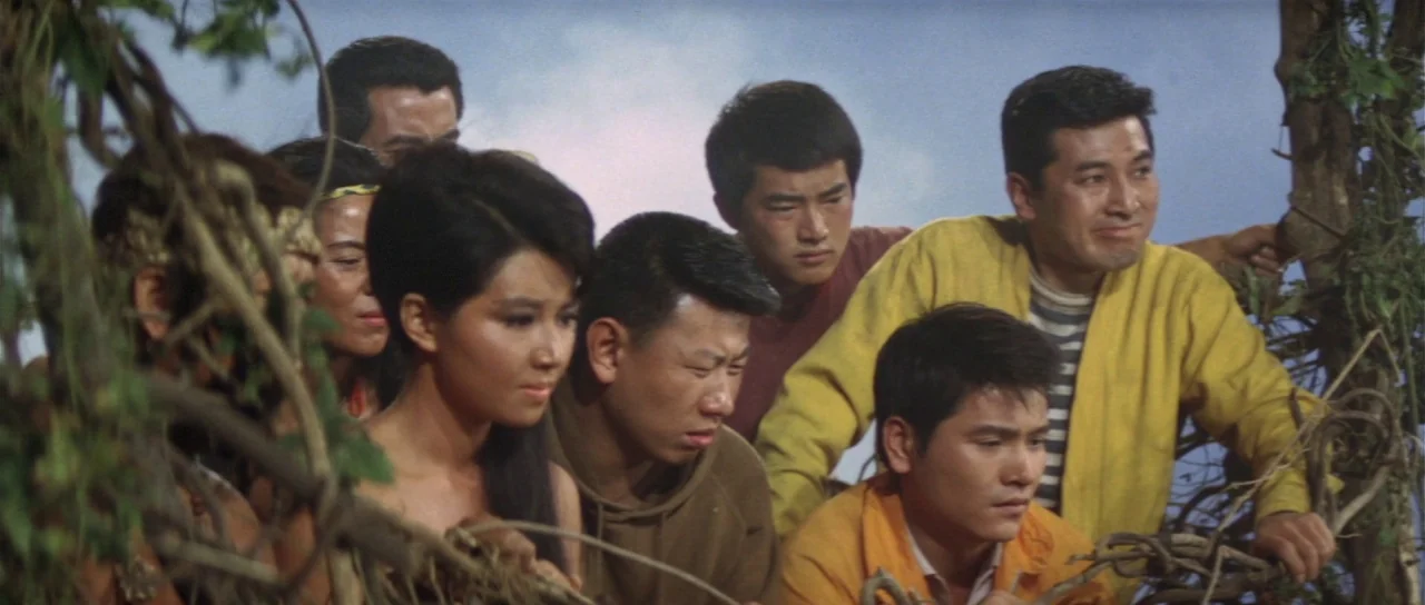 Ebirah Horror of the Deep (1966) movie still: medium — A group of people are huddled together outdoors near some thick tree branches. They look…