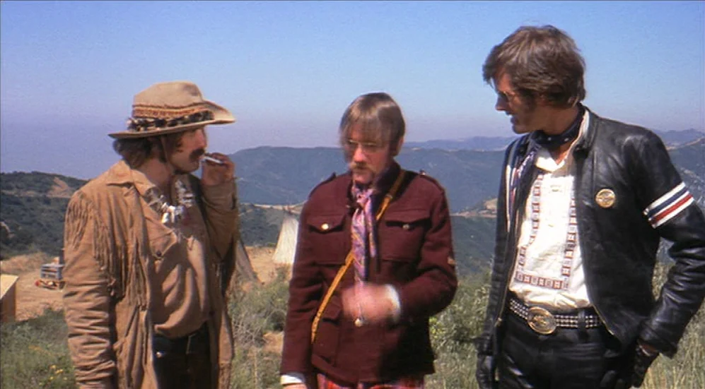 Easy Rider (1969) movie still: fullbody — Three men stand on a dry hillside under a cloudless blue sky. They are wearing varied 1960s attire while…