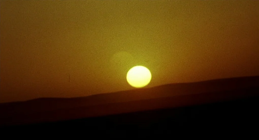 Furia (1999) movie still: establishing — A vast, barren desert landscape stretches out under a hazy, sunset-lit sky. The bright circular sun dips…