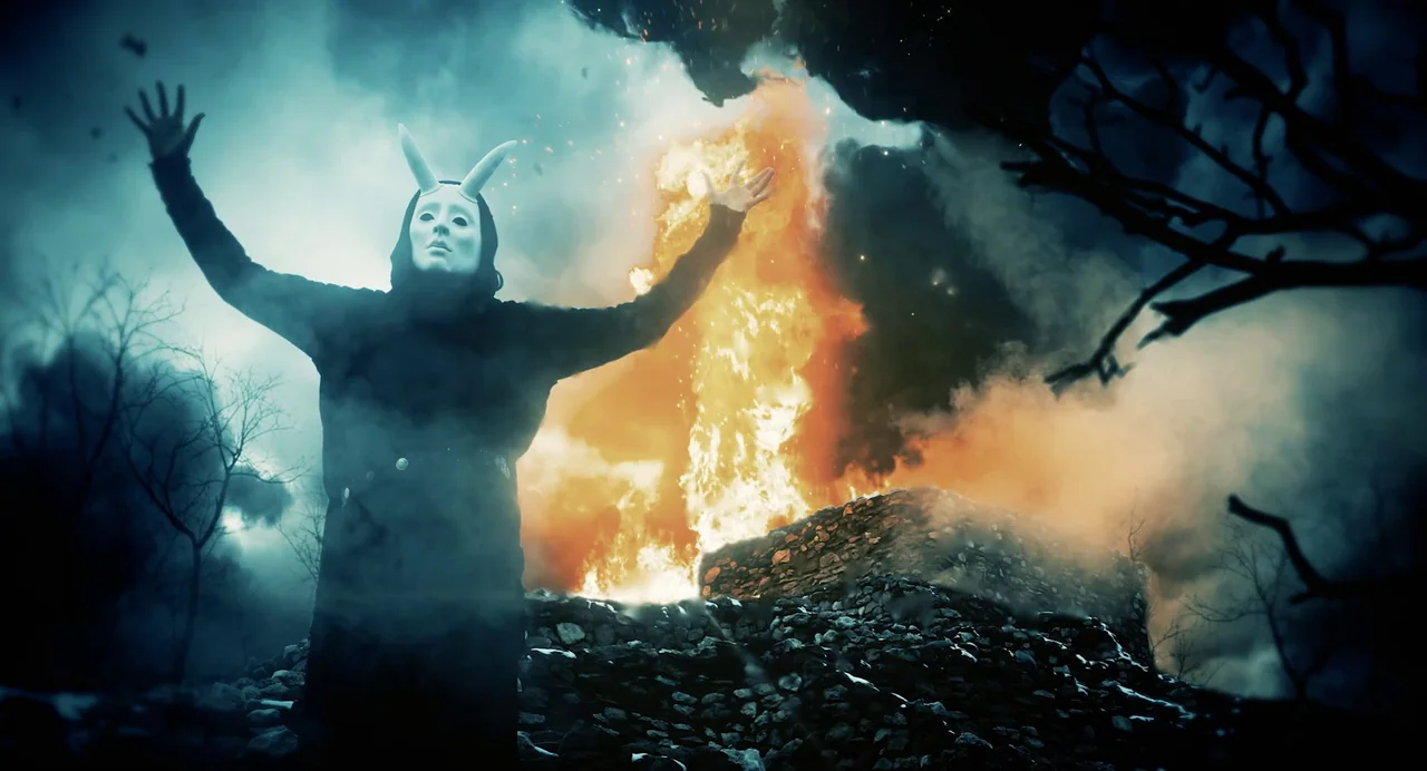Hellbender (2021) movie still: medium — A dark, smoky outdoor setting features a burning stone foundation beneath a stormy sky. A figure in a white…
