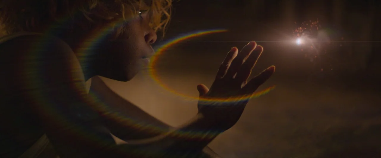The New Boy (2023) movie still: closeup — A dark, indistinct outdoor setting is illuminated by a small, bright point of light. A child with curly…