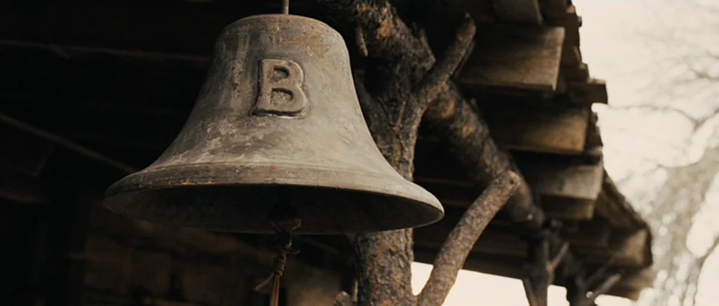 True Grit (2010) movie still: closeup — A weathered metal bell with the letter B cast onto its side hangs outside. It is suspended from a rustic…