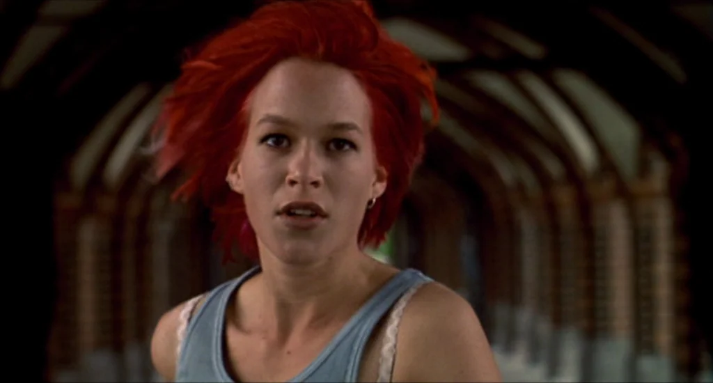 Run Lola Run (1998) movie still: medium — A stone vaulted passageway with repeating arches recedes into a darkened background. A young woman with…