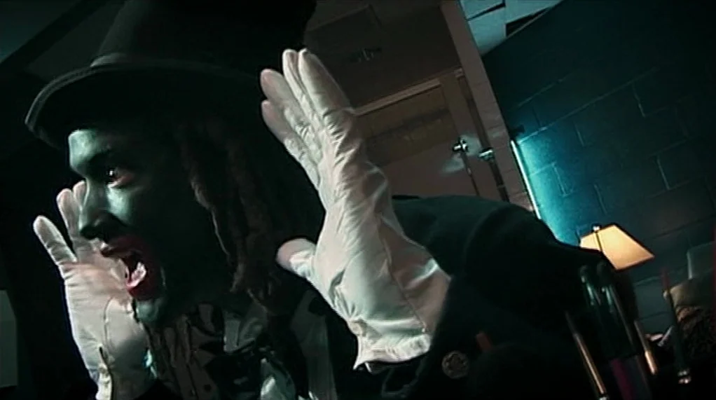 Bamboozled (2000) movie still: closeup — A dark, dimly lit space features a cinder block wall and a small lamp. A man in a top hat and green face…
