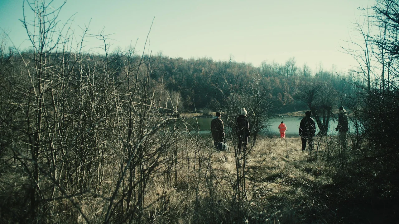 Helll Hole (2024) movie still: wide — A dry, grassy bank overlooks a calm lake backed by a dense deciduous forest. Four people are walking through…
