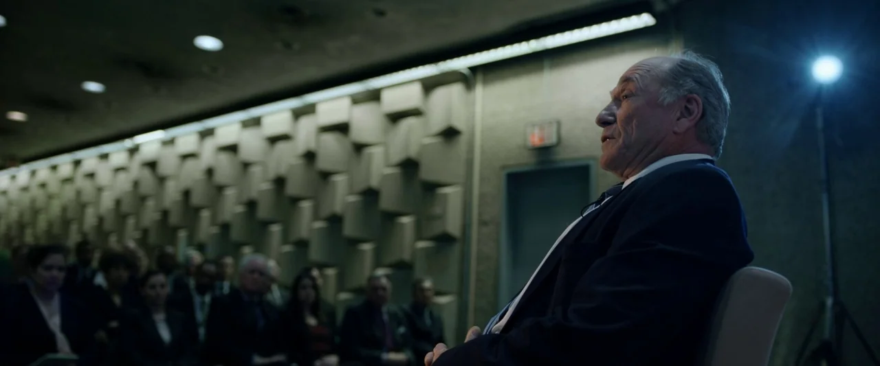 The Report (2019) movie still: medium — An interior space features a textured geometric wall panel with a crowd seated blurred. An older man sits in…