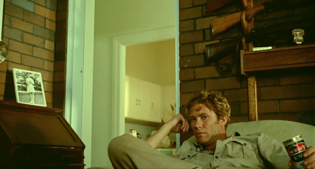 Wake In Fright (1971) movie still: medium — A residential room features dark brick walls adorned with rifles on a wooden rack. A young man relaxes…