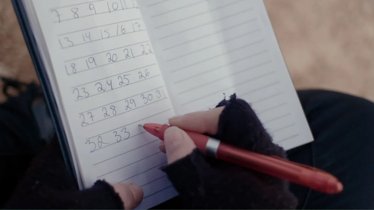 Code of Trust (2019) movie still: closeup — A notebook containing a list of numbers is held in a person's gloved hands. The person writes the number…