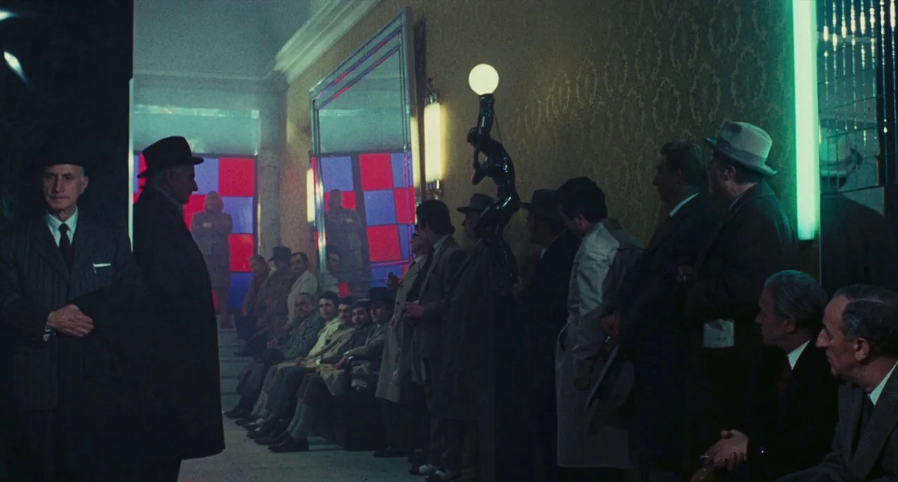 Roma (1972) movie still: wide — A crowded interior space features wallpapered walls with red checkered glass panes at the end. Men in suits and hats…