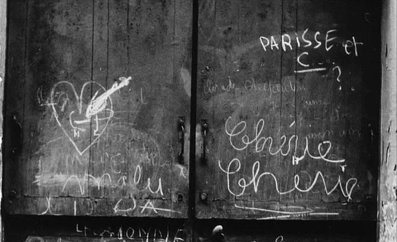 La Jetée (1962) movie still: closeup — A weathered wooden door features vertical panels and simple dark metal handles. Scratched graffiti markings,…