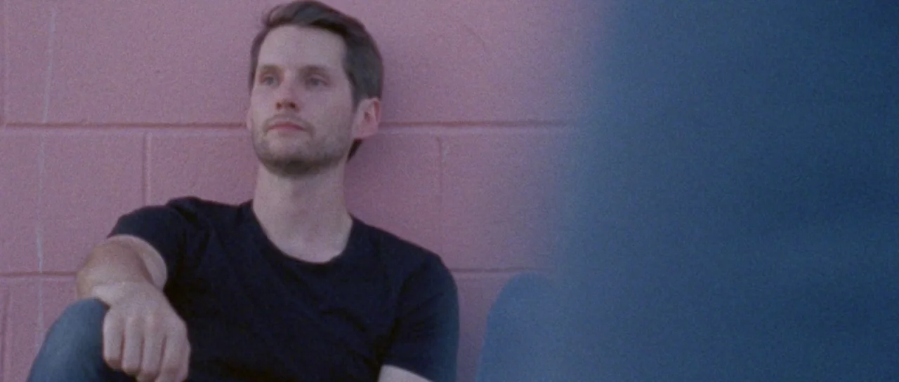 Starfish (2018) movie still: medium — A man sits against a textured pink concrete block wall outdoors. He looks forward with a neutral expression…