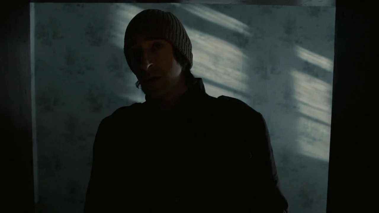 Splice (2009) movie still: medium — A man stands in a dark doorway against a wall with faded patterned wallpaper. Slanted light streaks across the…