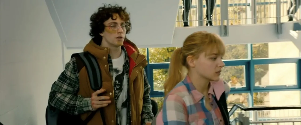 Kick-Ass 2 (2013) movie still: medium — A school hallway features a blue-framed glass wall and an industrial metal staircase railing. A male student…