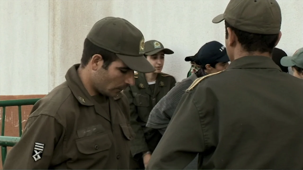 Offside (2015) movie still: medium — An outdoor area features a plain wall and military personnel standing in uniform. Officers in olive green…