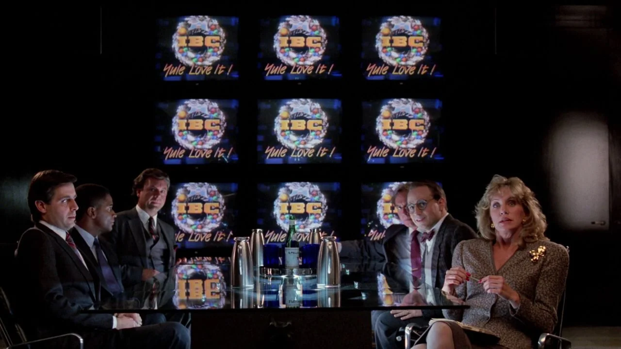 Scrooged (1988) movie still: wide — A dark boardroom is illuminated by a grid of nine glowing television screens. Five colleagues sit at a glass…