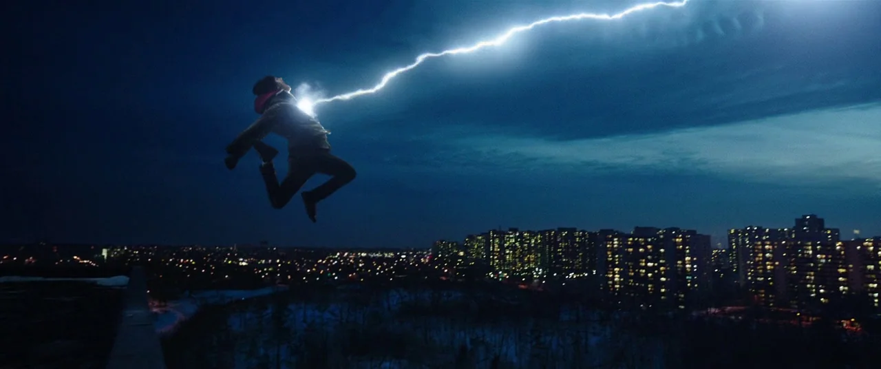 Shazam! (2019) movie still: wide — A night landscape features a glowing city with residential high-rises under a dark sky. A person leaps through…