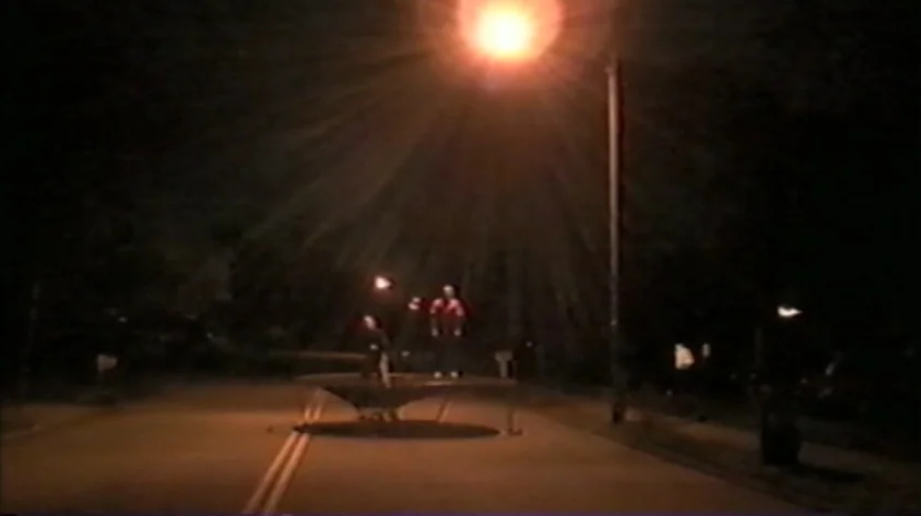 Trash Humpers (2009) movie still: wide — A paved residential road at night features a large trampoline placed in the center. Two individuals stand…