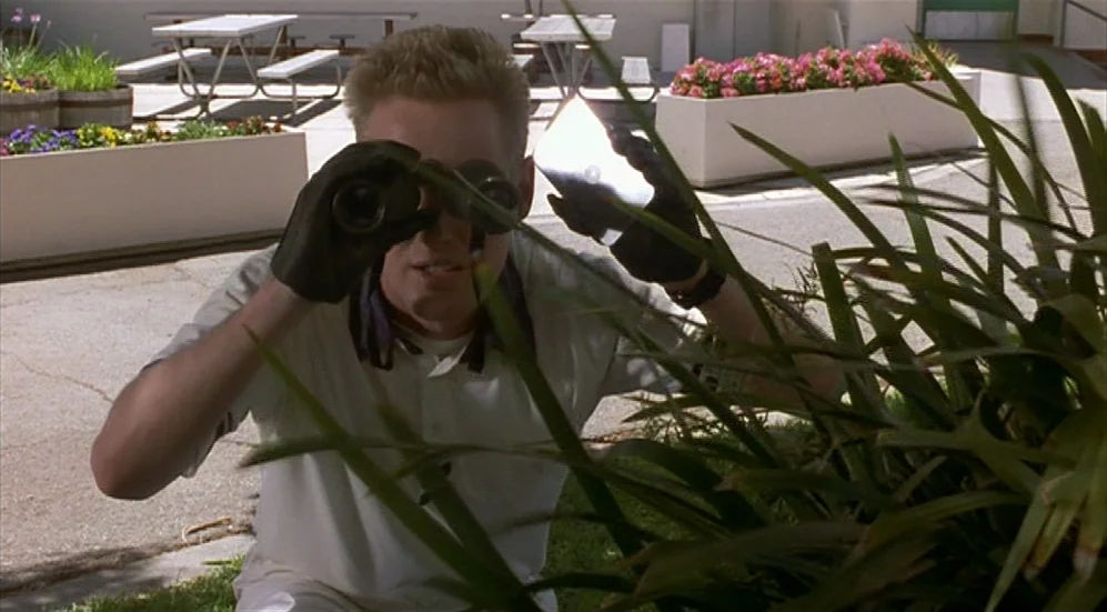 Bottle Rocket (1996) movie still: medium — A man crouches behind a leafy bush in a paved area with picnic tables. He holds black binoculars to his…