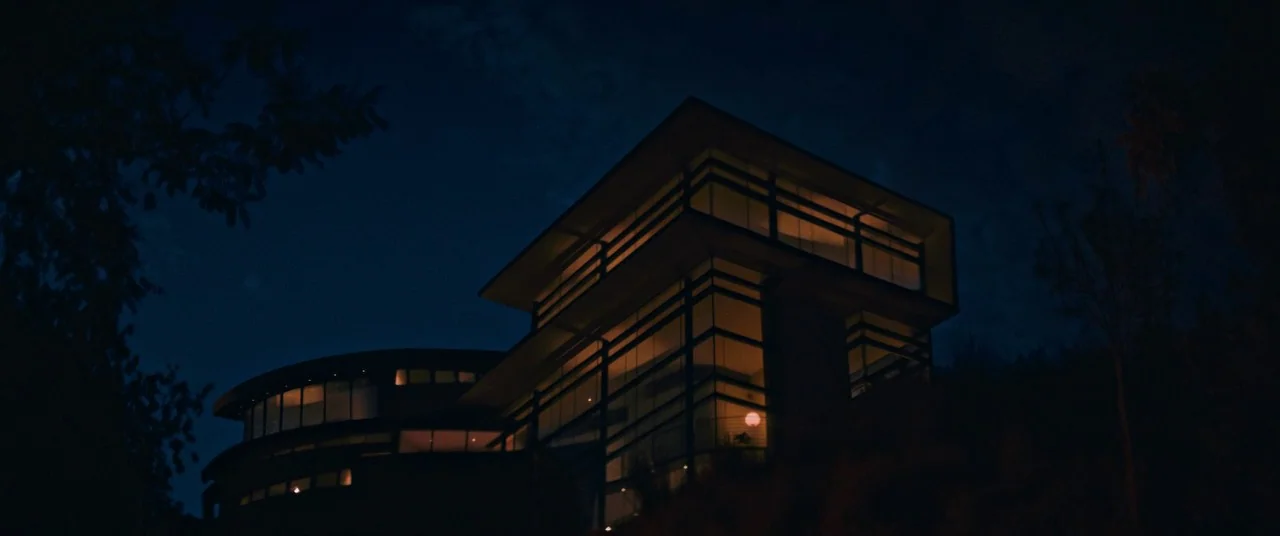 Elizabeth Harvest movie still: establishing — A modern multi-level mansion with floor-to-ceiling glass windows stands on a dark hill at night. Warm…