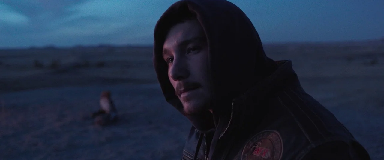 The Rider (2017) movie still: medium — A vast, empty desert expanse extends toward a dim horizon under a twilight sky. A man in a dark hooded jacket…