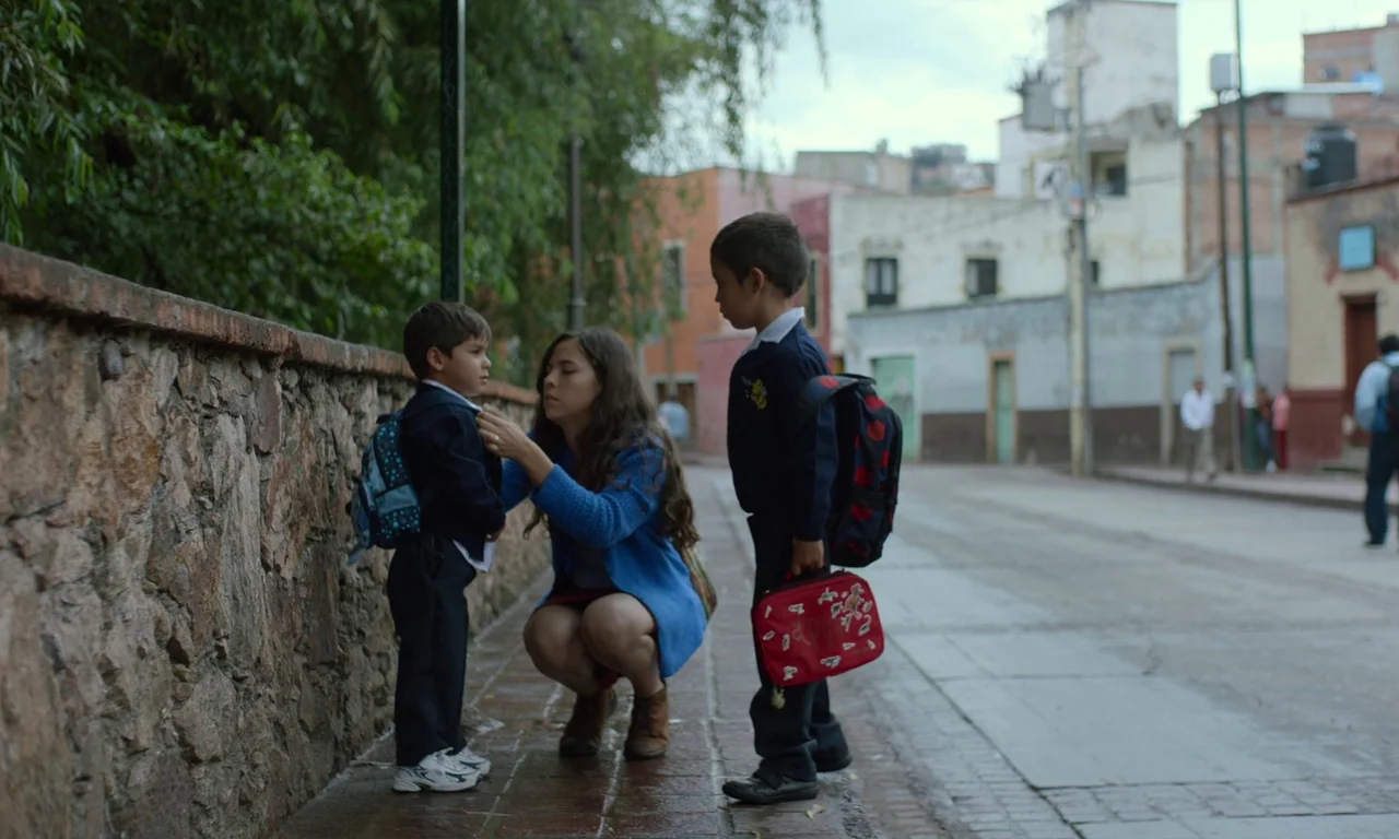 The Untamed (2016) movie still: fullbody — A quiet urban street lined with a rustic stone wall and low buildings. A woman crouches on the sidewalk…