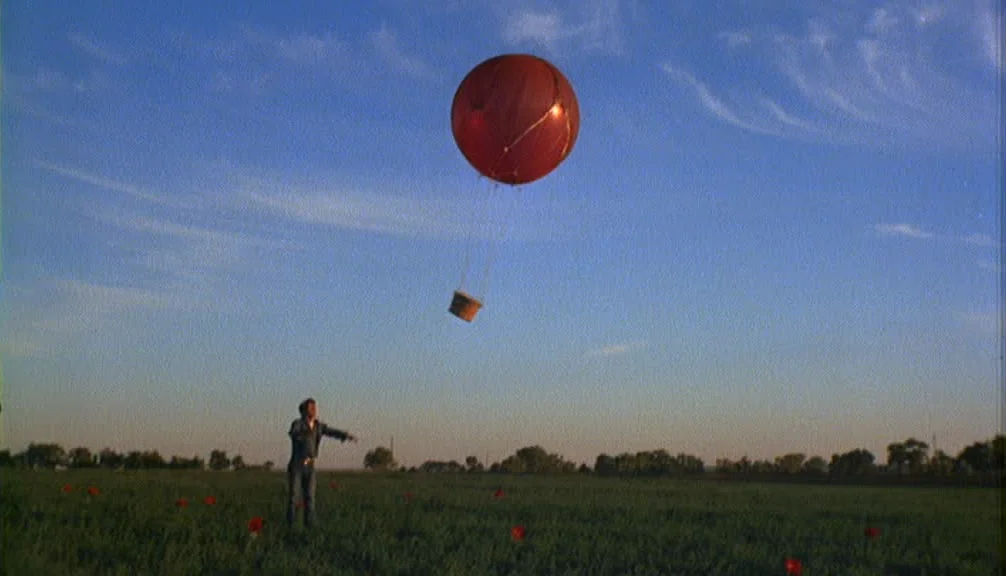 Badlands (1973) movie still: wide — A wide grassy field under a vast blue sky with light wispy clouds. A man stands alone reaching towards a small…
