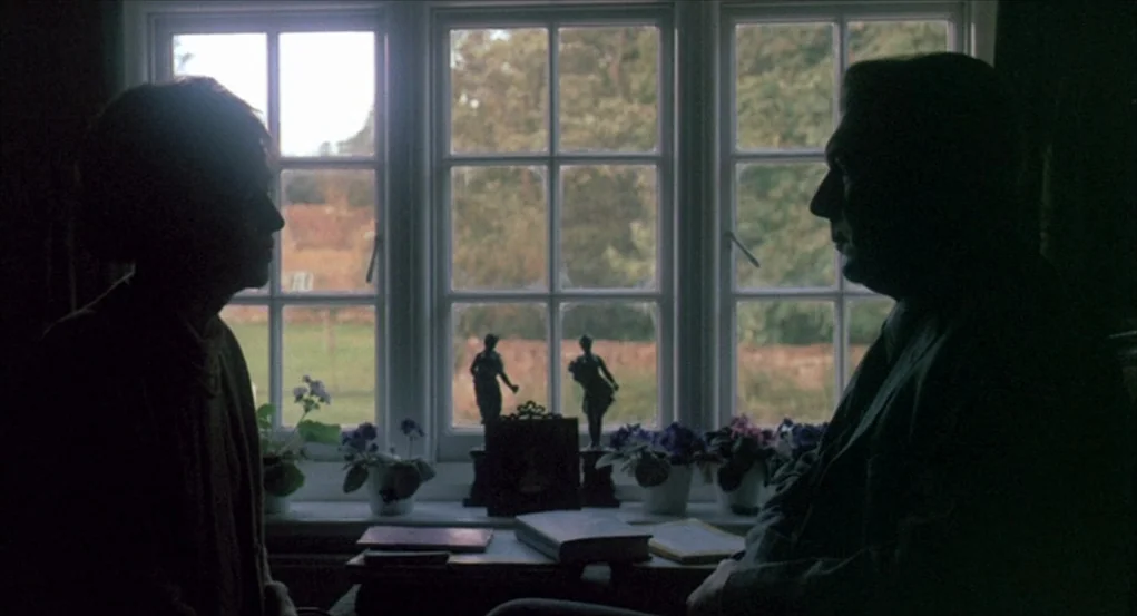 The Go-Between (1971) movie still: medium — A window with multiple panes overlooks a grassy lawn in the daytime. Two people in silhouette sit facing…
