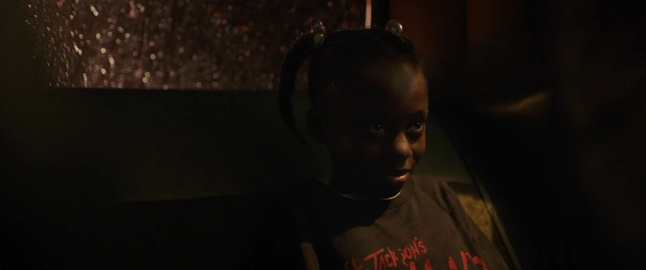 Us (2019) movie still: medium — A dark vehicle interior with a rain-slicked window visible in the background. A young girl with pigtails sits…