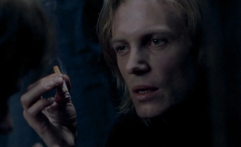 Pola X (1999) movie still: closeup — A man stands in a dark, poorly lit room with neutral colored surroundings. He holds a burning cigarette in his…