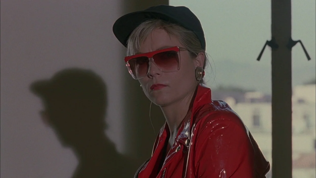Whore (1991) movie still: closeup — An interior space with a window admitting bright daylight beside the subject. A woman in a red vinyl jacket and…