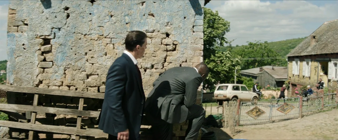 Heads of State (2025) movie still: fullbody — Two men in suits stand near a weathered blue stone wall in a rural area. The man in the grey suit…