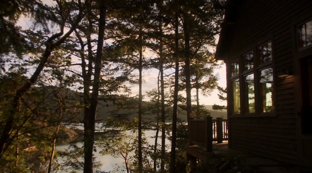 Your Sister’s Sister (2011) movie still: establishing — A wooden cabin sits at the edge of a tree-lined lake during sunset. Tall evergreen trees…
