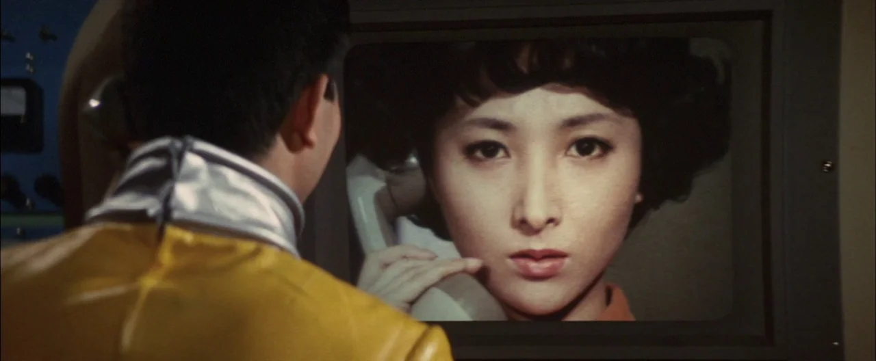 Destroy All Monsters (1968) movie still: medium — A dark, utilitarian room contains a large television screen displaying a woman on a call. The man…