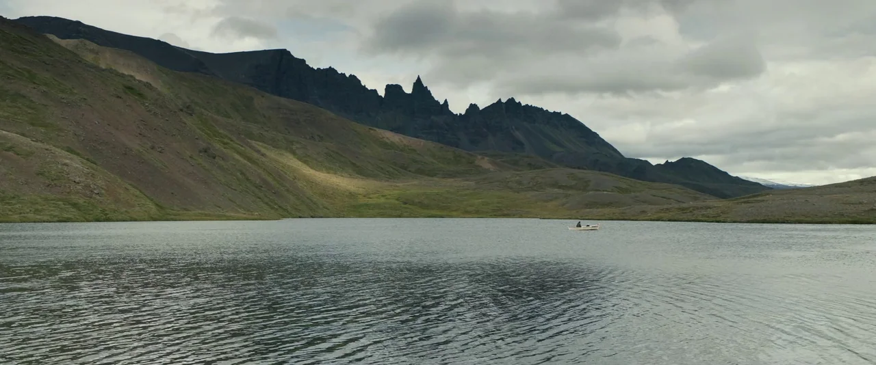 Babel (2006) movie still: wide — A wide, expansive mountain lake sits under a heavy, overcast cloudy sky. A solitary person drifts in a small white…