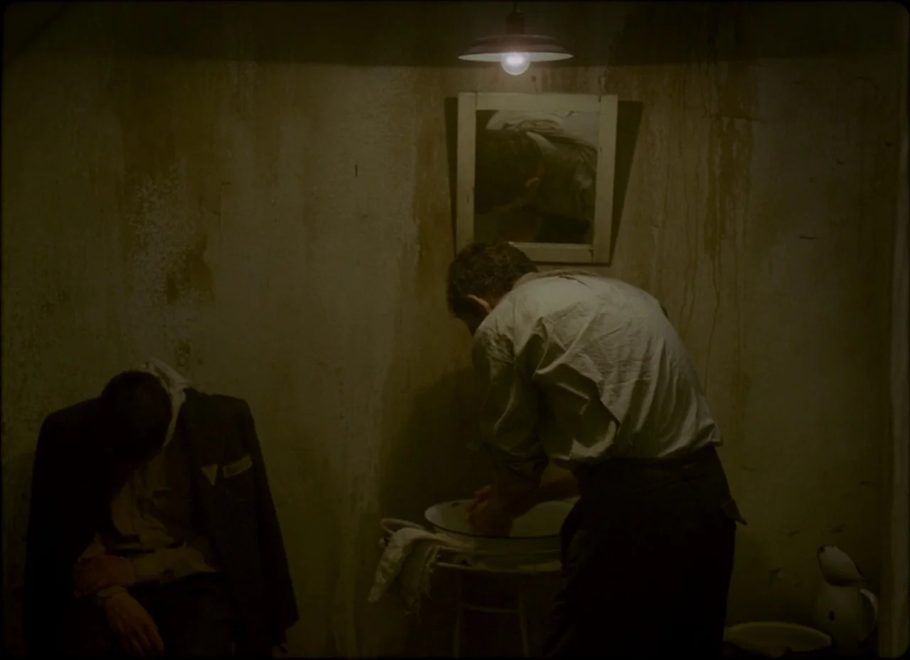 Son of Saul (2015) movie still: medium — A cramped, dimly lit room with peeling walls and visible stains under a bare bulb. A man stands washing his…