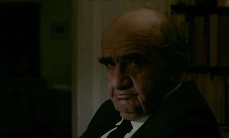 No End (1985) movie still: closeup — A dark study contains a bookshelf visible in the background shadow. An elderly man in a suit looks directly…