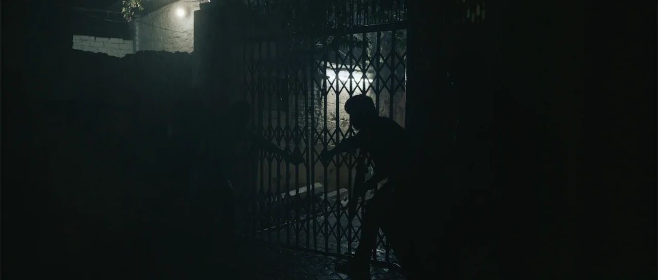 Nishaanchi 2 (2025) movie still: wide — A night scene shows a metal security gate blocking a dark alleyway entrance. Two silhouetted figures stand…