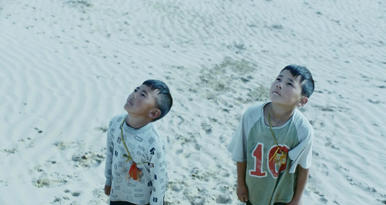 Balloon (2019) movie still: medium — Two young boys stand on a vast expanse of bright white sand. They tilt their heads back to look at something…