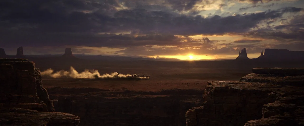 Rango (2011) movie still: establishing — A vast desert landscape features towering rock buttes silhouetted against a setting sun. A stagecoach moves…
